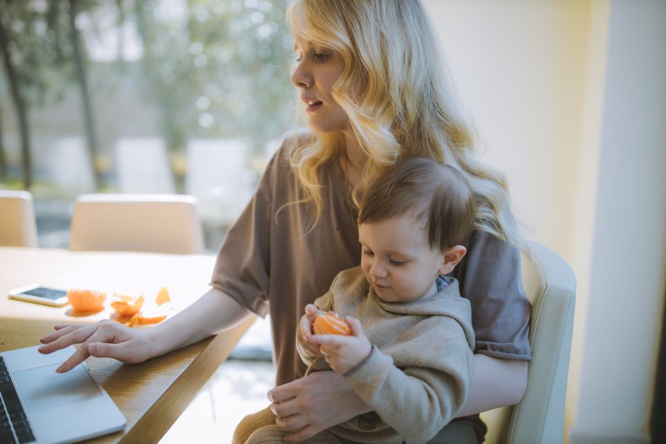 Mom working on laptop holding toddler eating orange