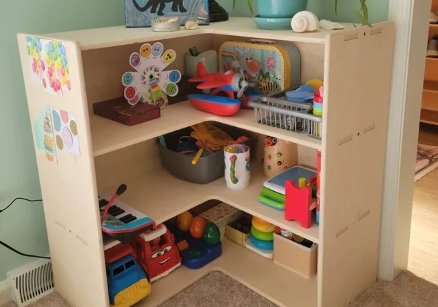 Light-coloured wooden toy display shelf situated in corner of living room and filled with colourful toys.