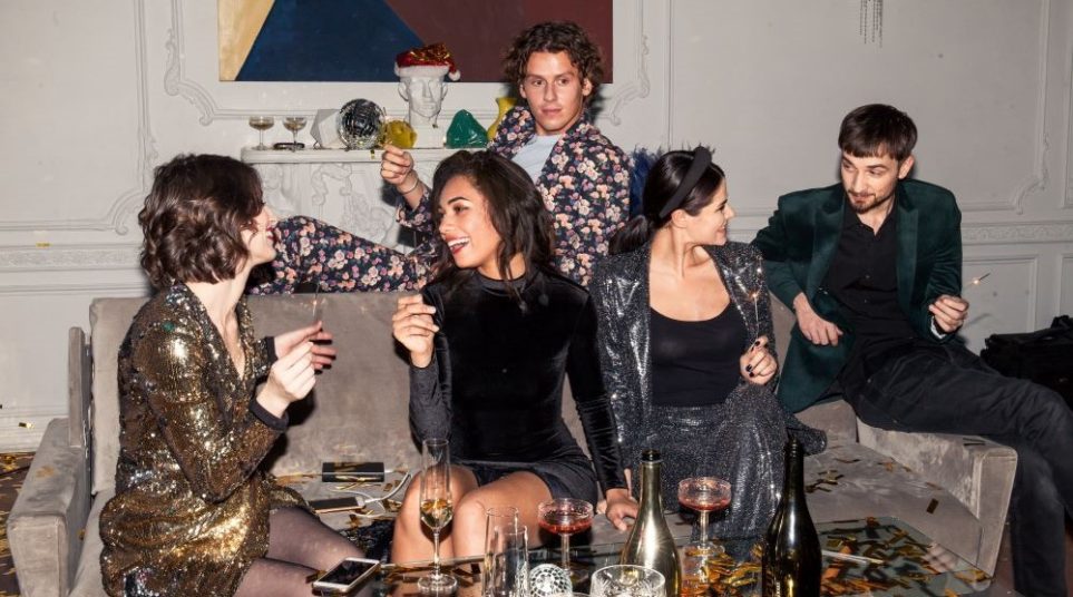 Five friends sit on a couch (with one young man sprawled across the top, wearing a floral print suit) smiling and socializing together while holding sparklers, with lots of metallic gold confetti scattered on the coffee table as well as champagne flutes and bottles