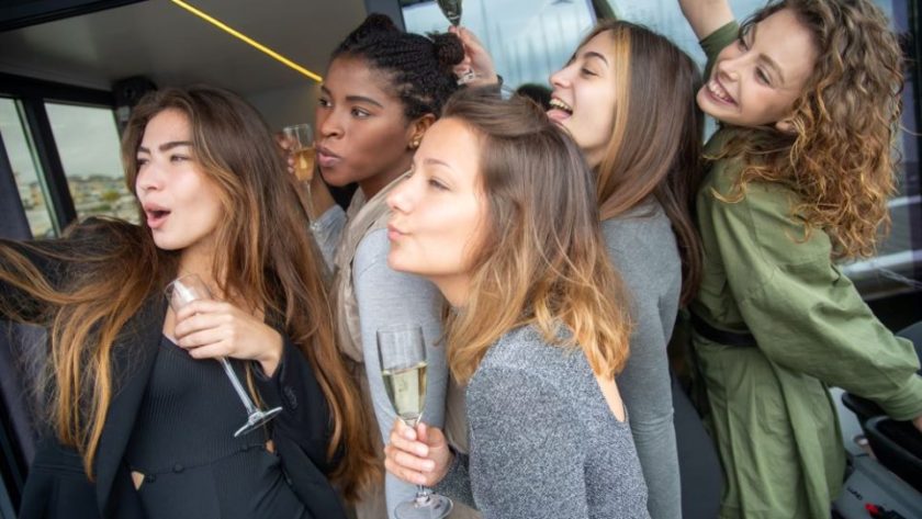 Five young women crowd together while smiling and posing for a camera to the left (out of sight), while holding champagne flutes