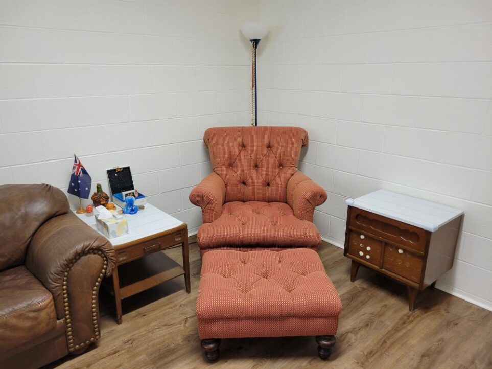 A basement corner furnished with a floor lamp standing behind an overstuffed red clay/orange-coloured armchair and matching footstool. To the right is a small end table and to the left is another small end table covered in some decorations. To the left of that is a brown leather couch (mostly out of picture). When you furnish a house, it's important to create inviting seating areas even in underused spaces like basements