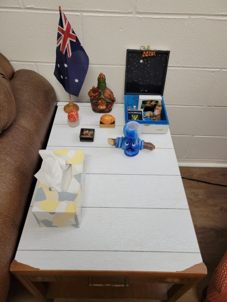 The end table on one side of the brown couch in the basement is topped with shiplap peel-and-stick wallpaper and features a tissue box plus older nostalgic decor including a miniature Australian flag, trinkets from Costa Rica, Russia, Germany, and other gifted keepsakes.