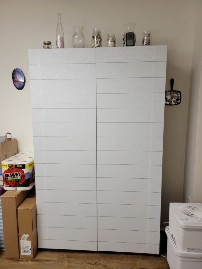 A brown cabinet covered in shiplap peel-and-stick wallpaper is positioned against a basement wall next to a dehumidifier on one side and a pile of small cardboard boxes and paper towel package on other side. On top are several decorative bottles and clear glass vases filled with seashells. To each side, hanging on the wall, is a coastal decoration