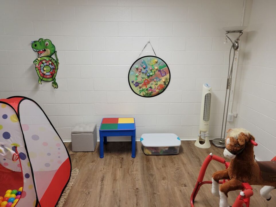 Back area of basement: a floor lamp stands in one corner next to a fan. A small Lego table and a couple of storage bins of toys line the back wall, with two kids Velcro 'dart' boards hanging on the wall. In partial view to the left is one of the boys' play tents, and to the right is a ride-on pony (toy overflow area)