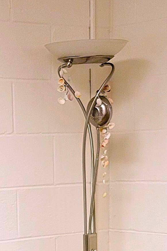 A metal floor lamp stands in an otherwise empty basement corner. From it hang small seashells strung on fishing line for a decorative effect