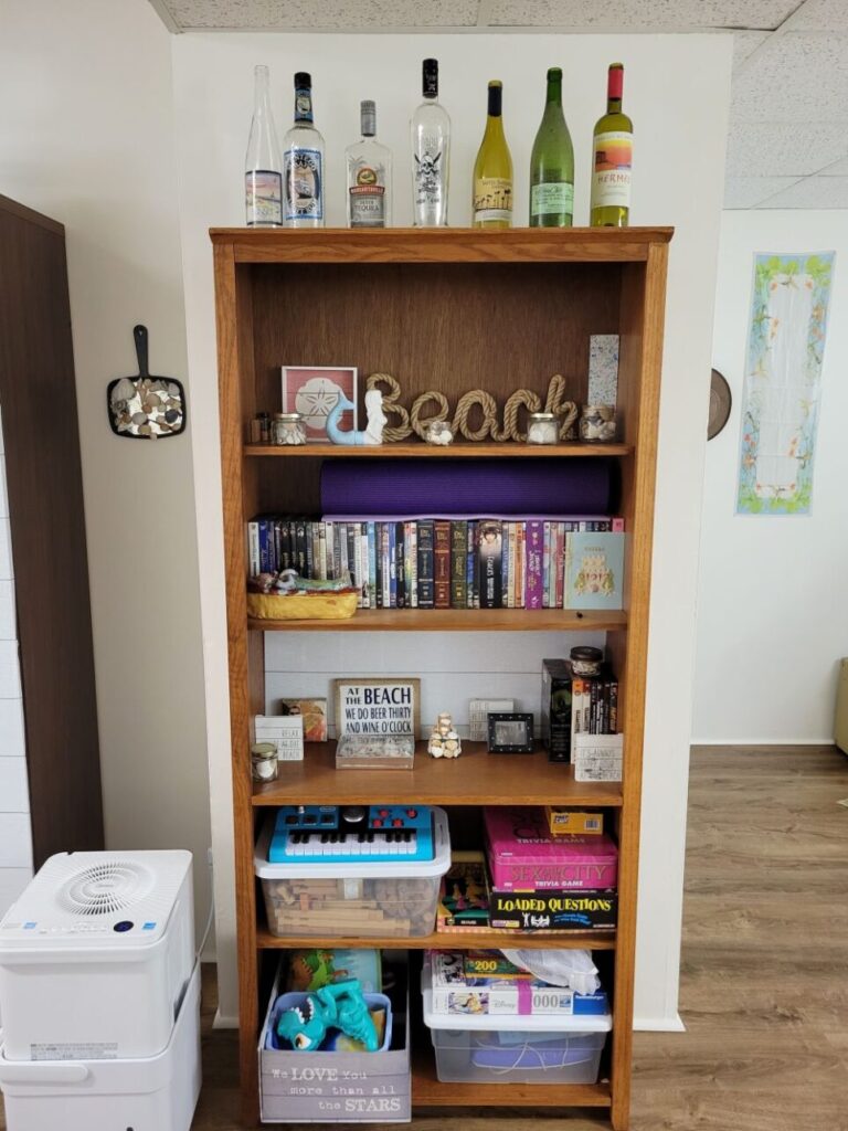 A brown bookcase is positioned against a basement wall with a dehumidifier on one side and an open space on the other side, each side with decorations hanging on the wall. Atop the bookcase are several nautical-themed wine and liquor bottles, and on the top shelf is coastal decor ('beach' spelled out in cursive in nautical rope, a ceramic mermaid, small jars of seashells, etc.). On lower shelves are DVDs, more beach decor, board games, and toys. Mixing familiar, preferred aesthetics and nostalgic personal items is how to make a house feel like a home