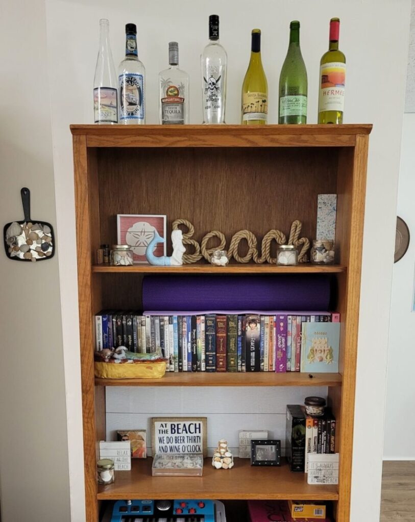 A brown bookcase (only upper half shown) features coastal-themed decorations on two of the shelves plus DVDs and games on other shelves. On top are nautical-themed wine and liquor bottles. To the left side is an old broken mirror with jagged pieces of glass and seashells glued on as a coastal art-piece. Furnish a house with beloved decor even in underutilized spaces like the basement, so that every area feels inviting.