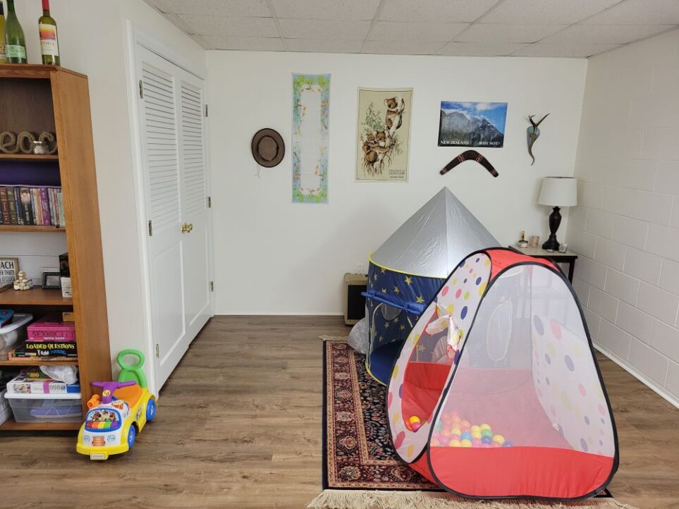 The back open area of the basement: two colourful play tents are positioned on top of the medium-sized area rug in contrast to the demure colours and relaxed decor of wall decorations and corner table with lamp and small decor. Another colourful small ride-on toy is 'parked' to the left, next to the brown bookcase.