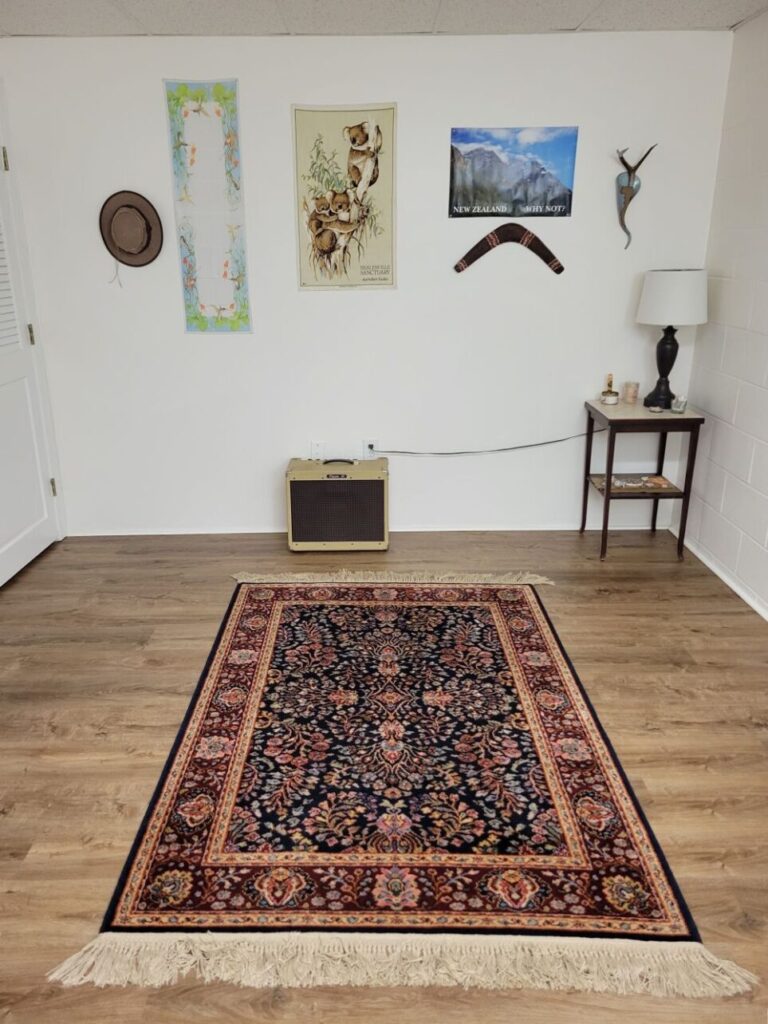 A medium-sized Persian-style area rug lays in the middle of the open area at the end of the basement room. In one corner part of one of the closed doors to the furnace is seen and in the other corner is a small end table with a lamp on top along with small decorations. An old amplifier sits against the wall as the only other standing item. On the wall are hung several travel-inspiring decorations and keepsakes from around the world - an Aboriginal boomerang, a New Zealand poster with a mountainscape, a koala cloth wall hanging, and more.