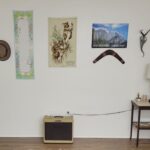 The back wall of the basement room is lined with only a vintage amplifier and a small brown end table in the corner with a brown lamp on top as well as small coastal and natural decor items. Several decorations hang from the wall including a hat with an emu feather, boomerang, and cloth koala hanging from Australia, a decorative heirloom scarf, and other decor