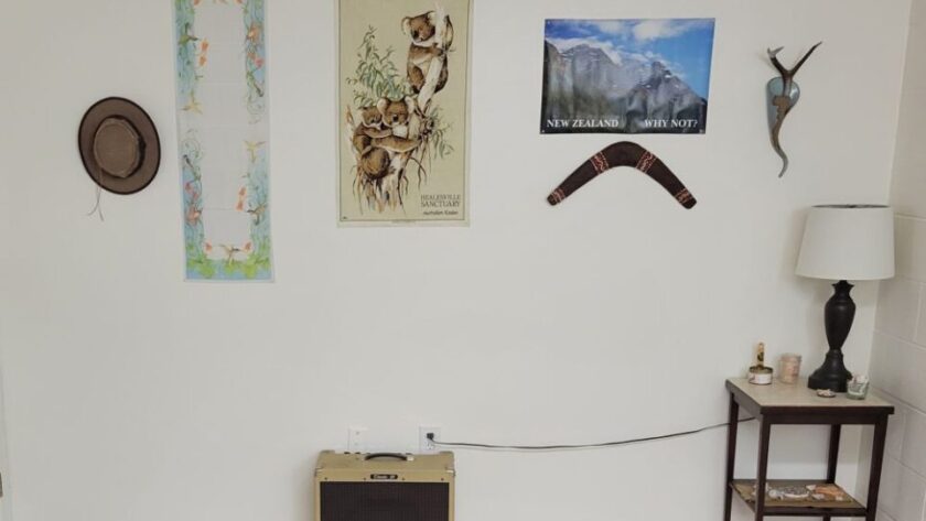 The back wall of the basement room is lined with only a vintage amplifier and a small brown end table in the corner with a brown lamp on top as well as small coastal and natural decor items. Several decorations hang from the wall including a hat with an emu feather, boomerang, and cloth koala hanging from Australia, a decorative heirloom scarf, and other decor