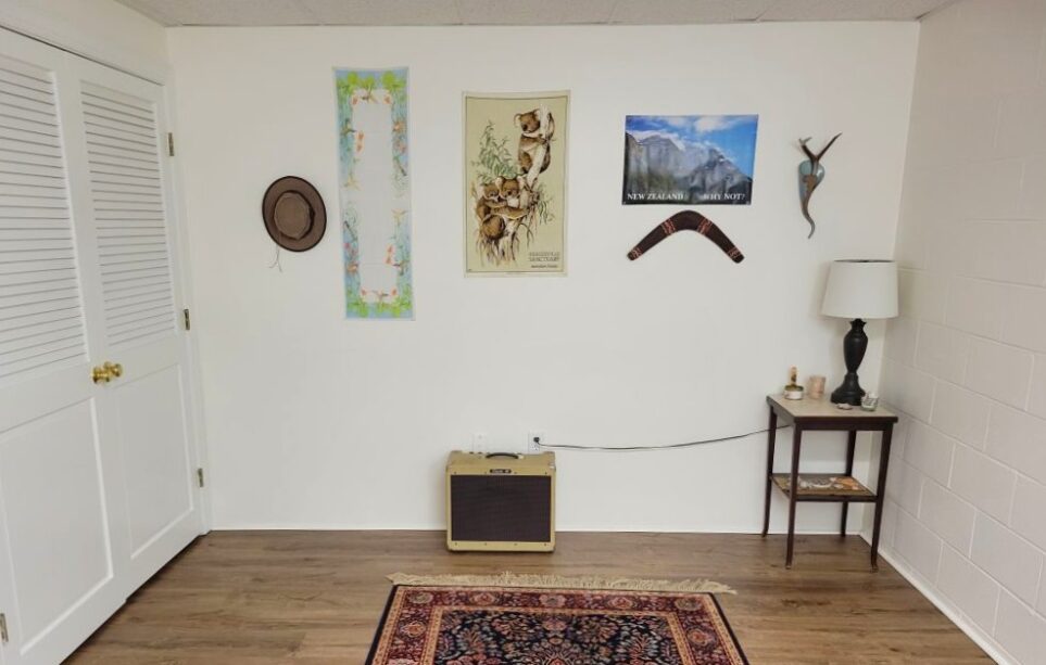 The back area of the basement room: a medium-sized Persian-style area rug sits in front of the back wall lined with only a vintage amplifier and a small brown end table in the corner with a brown lamp on top as well as small coastal and natural decor items. Several decorations from or featuring other areas of the world hang on the wall, including souvenirs from Australia, a decorative heirloom scarf, and other decor. When you furnish a house, give care and attention to even the most remote corners - including the back of the basement, to make them happy spaces.