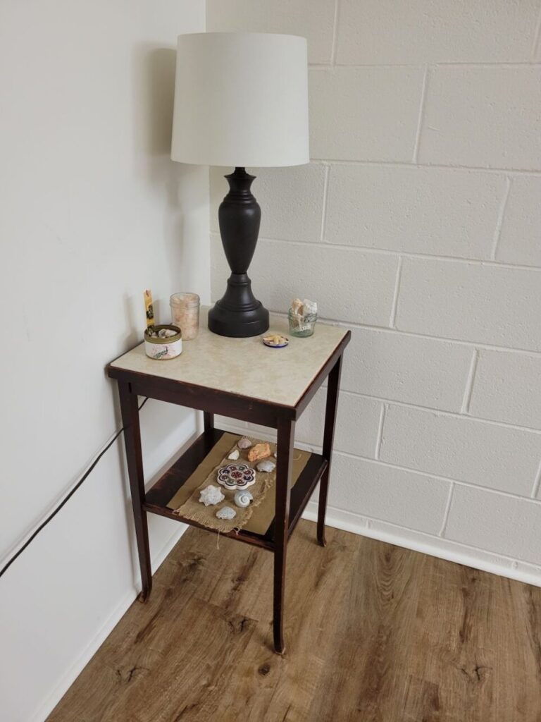 In a basement corner is a small end table with a table lamp and decorative items on top, as well as on the lower shelf, including small decorative jars of seashells and other natural relics