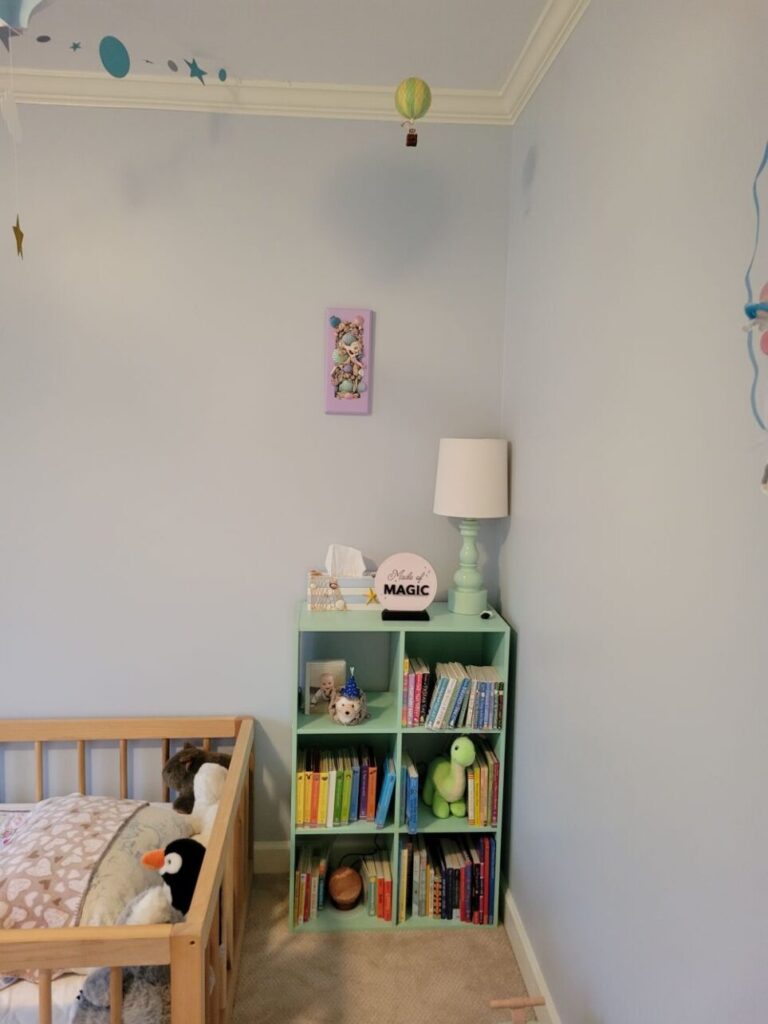 In the corner of the baby's room is a sea green cubby shelving unit with a sea green lamp on top next to a decorative tissue box and other decor. Board books line the shelves alongside a few small plush animals. To the left of the shelves (and mostly out of shot) is a floor bed made up with plush animals at the end and soft blankets