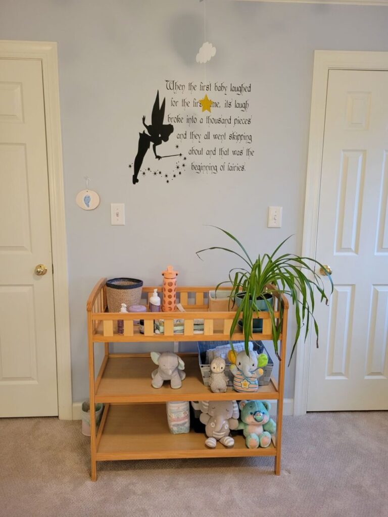 Above the changing table in the baby's room is a Peter Pan quote decal on the wall which reads: 'when the first baby laughed for the first time, its laugh broke into a thousand pieces and they all went skipping about and that was the beginning of fairies.' A silhouette of Tinkerbell flanks the left side of the text. On the top shelf of the changing table is a healthy airplane plant next to necessities such as water, lotion and pull-ups. On lower shelves are plush animals and extra pull-ups. To either side is a door (one leading into the closet and one leading out of the bedroom and into the hallway). This is how to make a house feel like a home: I've decorated it in the same way as the previous home, from buying the same wall decal to showcasing the same beautiful plant on the same changing table