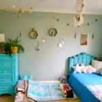 The children's bedroom (technically the kindergartener's room but where both boys now sleep at night) is furnished with a twin bed and blue tufted headboard on the right, covered in star-shaped throw pillows among other pillows and a blue blanket. On the left is a blue-painted dresser with an airplane plant, lamp, and other decor on top. On the floor in between the twin bed and dresser is a twin futon mattress tucked partially under the twin bed where the toddler sleeps, and is furnished with plush animals, several little pillows, and matching blanket. From the ceiling hangs a dreamcatcher in the center, a fishnet from the corner with nautical relics caught in it, and other nautical decor on the wall.