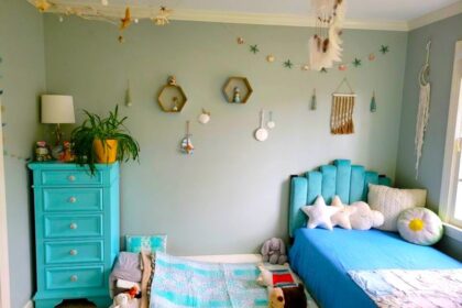 The children's bedroom (technically the kindergartener's room but where both boys now sleep at night) is furnished with a twin bed and blue tufted headboard on the right, covered in star-shaped throw pillows among other pillows and a blue blanket. On the left is a blue-painted dresser with an airplane plant, lamp, and other decor on top. On the floor in between the twin bed and dresser is a twin futon mattress tucked partially under the twin bed where the toddler sleeps, and is furnished with plush animals, several little pillows, and matching blanket. From the ceiling hangs a dreamcatcher in the center, a fishnet from the corner with nautical relics caught in it, and other nautical decor on the wall.