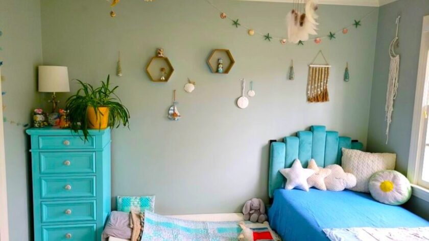 The children's bedroom (technically the kindergartener's room but where both boys now sleep at night) is furnished with a twin bed and blue tufted headboard on the right, covered in star-shaped throw pillows among other pillows and a blue blanket. On the left is a blue-painted dresser with an airplane plant, lamp, and other decor on top. On the floor in between the twin bed and dresser is a twin futon mattress tucked partially under the twin bed where the toddler sleeps, and is furnished with plush animals, several little pillows, and matching blanket. From the ceiling hangs a dreamcatcher in the center, a fishnet from the corner with nautical relics caught in it, and other nautical decor on the wall.