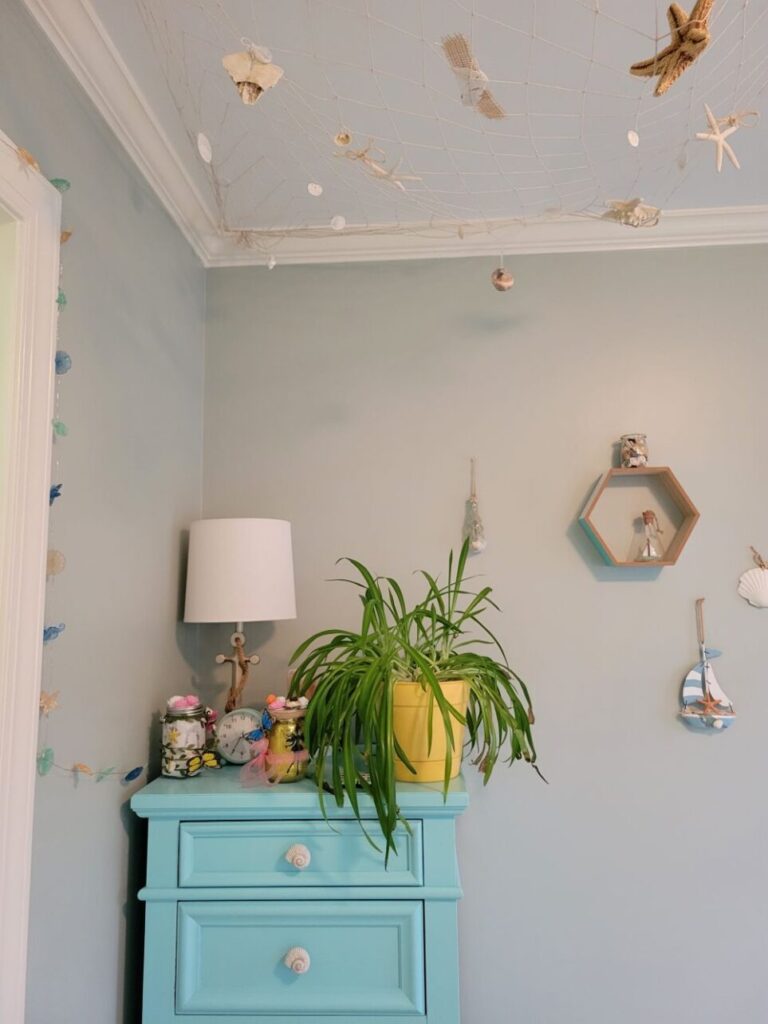In the corner of the kindergartener's room hangs decorative nautical fishnet from the ceiling with starfish, sand dollars and seashells stuck in it. A blue-painted dresser with seashell drawer knobs is positioned in the corner with an anchor lamp, airplane plant, and decorative items on top. Nautical decor hangs on thew all to the right and out of view.