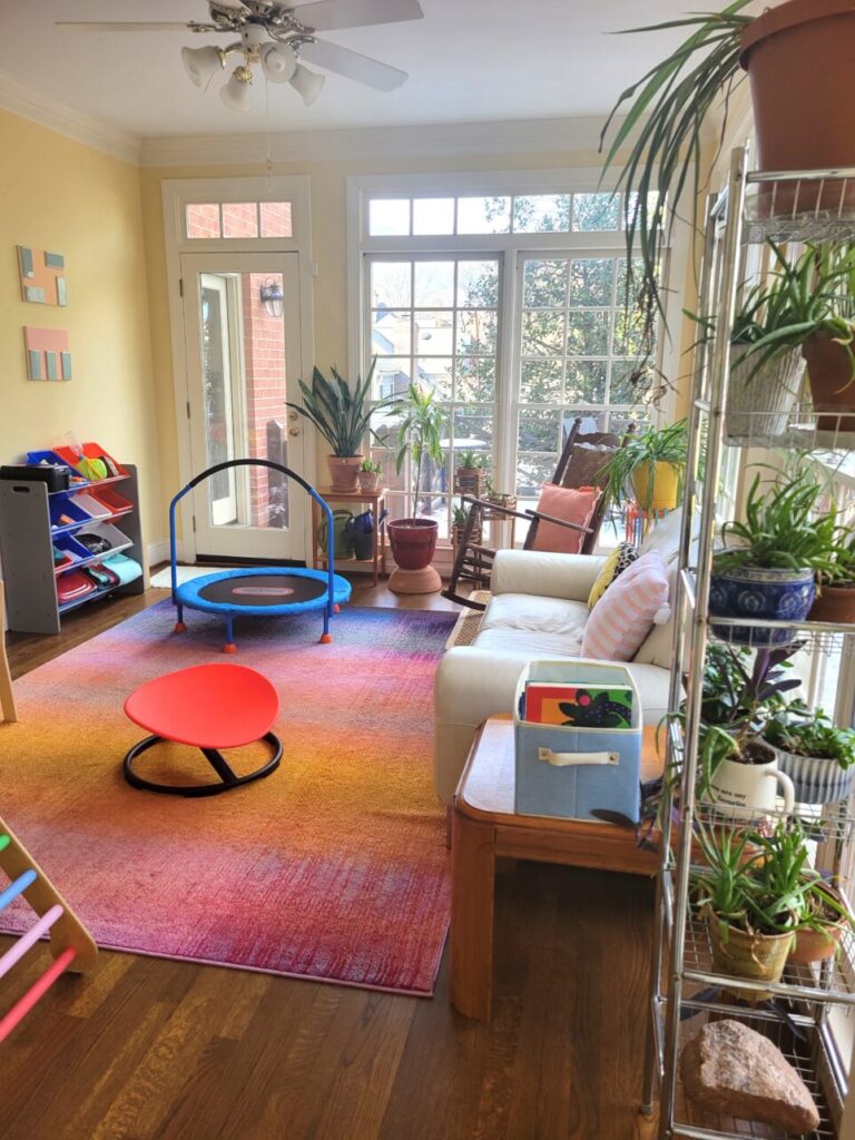 View of sunroom/playroom with Pikler triangle partially in view to left, mini trampoline toward back wall, several plants, rocking chair in far corner, and white loveseat to right along with tall open shelving unit with several succulent plants next to it