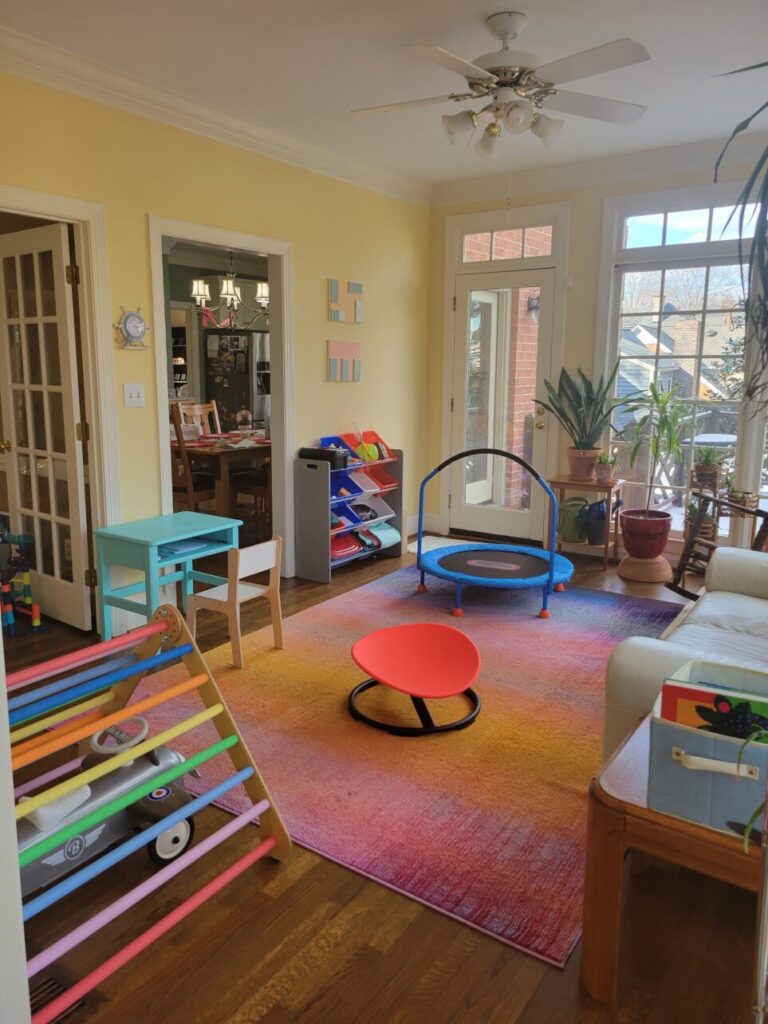 View of sunroom/playroom with Pikler triangle on left and ride-on airplane underneath, blue-painted children's desk against far wall with chair, mini trampoline toward back wall, several plants, and white loveseat partially out of sight to right