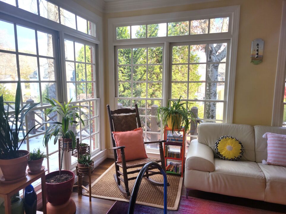 View of corner of sunroom with woven rattan square rug under heirloom rocking chair, several plants to left of that in front of second windowed wall, and a plant atop a colourfully-painted end table and white loveseat to right
