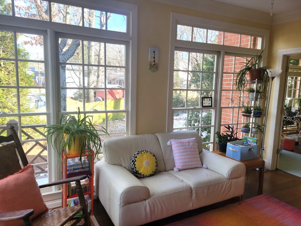 View of sunroom with window-filled wall behind white loveseat and colourful throw pillows, with end tables on either side with plants on top. To left and partially out of sight is rocking chair with coral throw pillow. To right of other end table is tall shelving unit with many plants, and floor lamp next to it. Partially out of view and to very right is opening into study