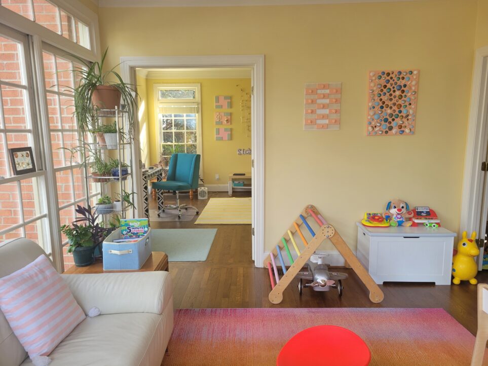 Creative spaces: the sunroom with a view of the study through the open French doors. Both with light yellow-painted walls, and a yellow/white striped rug seen in study along with a smaller sea green rug, and a colourful purple/red/orange rug in sunroom underneath bright red sensory spinning chair. Also in sunroom: Pikler triangle and other children's toys and toy box, and white loveseat (partially in view) next to end table with blue bin of books and many plants on open shelving unit next to it in front of windows. On wall above triangle and toybox are two shell collages with multi-colour painted seashells