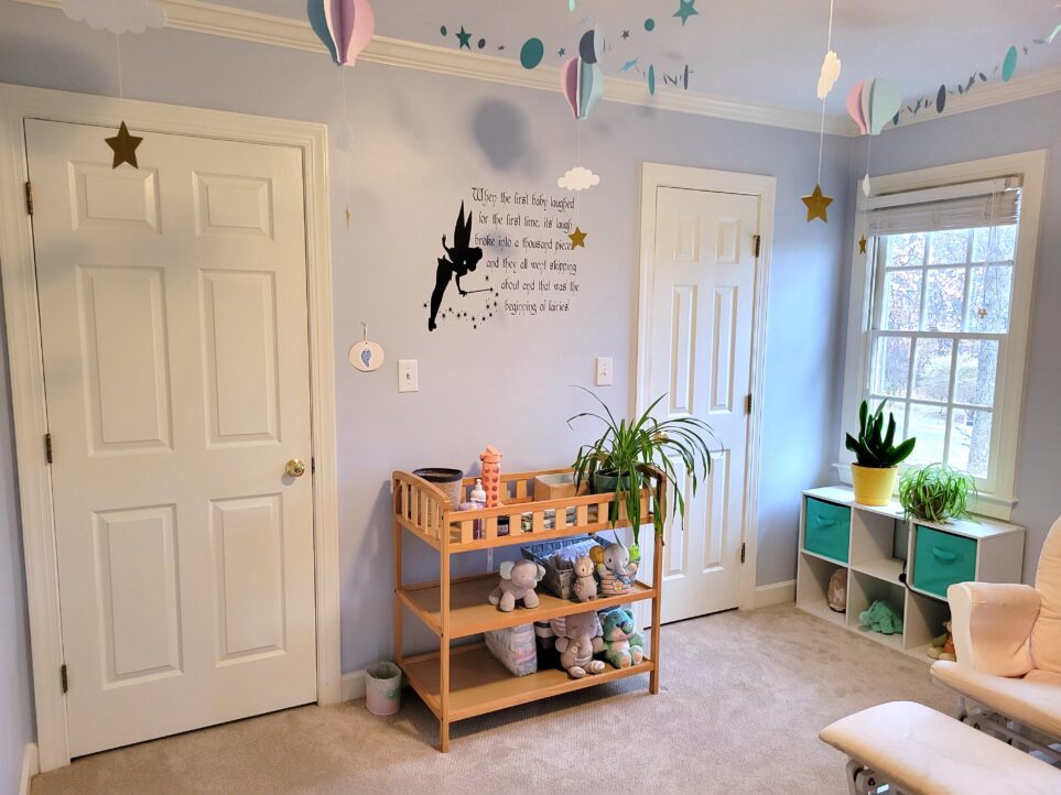 The baby's room: pastel-coloured paper cutout hot air balloons and stars hang from the ceiling, a Peter Pan fairies quote decal is affixed to wall above changing table that holds up healthy green airplane plant and other essentials like lotion, with cute plush animals resting on lower shelves. In the left corner is the bedroom door and in the opposite corner is the closet door next to a window above white cubby shelves, with two more plants on top and a salt lamp and more plush animals in lower shelves. To the right (and mostly out of sight) is a light pink glider rocker chair and footstool.