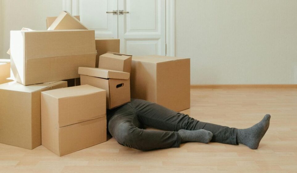 A person wearing gray socks and black jeans lays on the floor with their legs sticking out of a pile of empty moving boxes covering their entire upper half (someone has 'given up'). Giving yourself plenty of time to pack is a key component to how to make a house feel like a home, so that way the moving process isn't entirely stressful and rushed