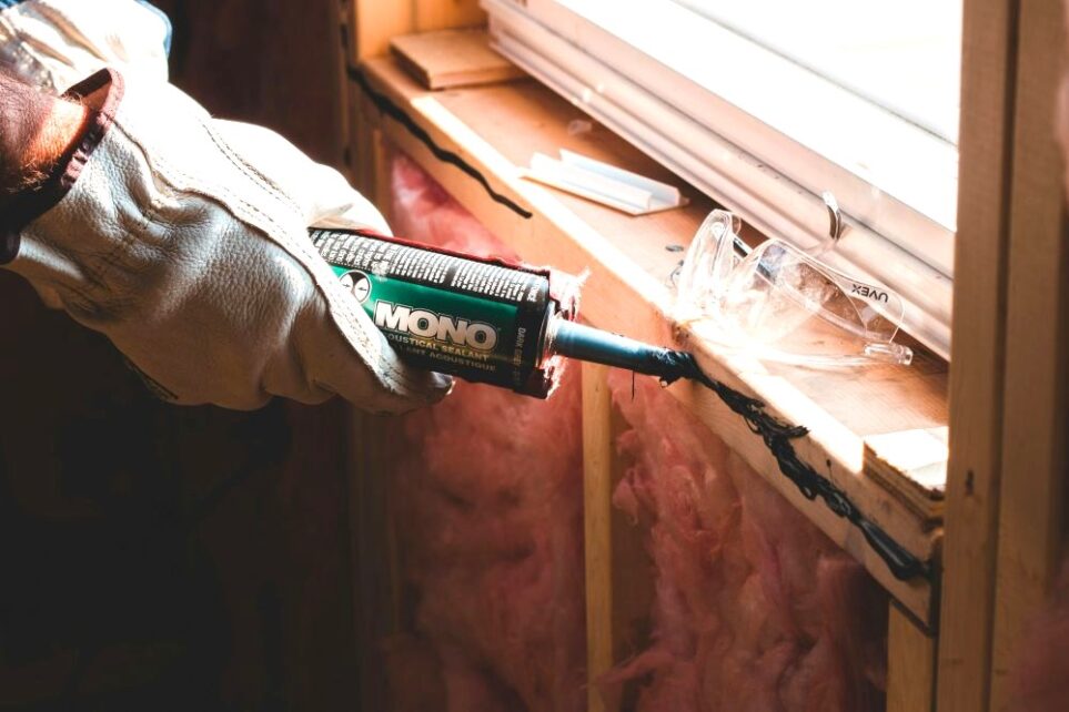 Close-up photo of gloved hands holding sealant and applying it at base of window sill in unfinished space