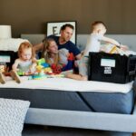 A couple and two young children sit on a bare king bed mattress with moving boxes positioned around them. A little girl plays with toys scattered around while the little boy pulls items out of one of the boxes. Parents lounge on bed between the children looking happily relaxed. This is how to make a house feel like a home: allow the time and space in the process to keep spirits high and relaxed
