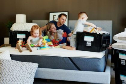 A couple and two young children sit on a bare king bed mattress with moving boxes positioned around them. A little girl plays with toys scattered around while the little boy pulls items out of one of the boxes. Parents lounge on bed between the children looking happily relaxed. This is how to make a house feel like a home: allow the time and space in the process to keep spirits high and relaxed