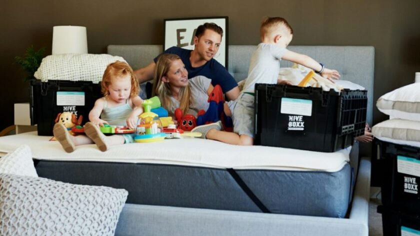 A couple and two young children sit on a bare king bed mattress with moving boxes positioned around them. A little girl plays with toys scattered around while the little boy pulls items out of one of the boxes. Parents lounge on bed between the children looking happily relaxed. This is how to make a house feel like a home: allow the time and space in the process to keep spirits high and relaxed