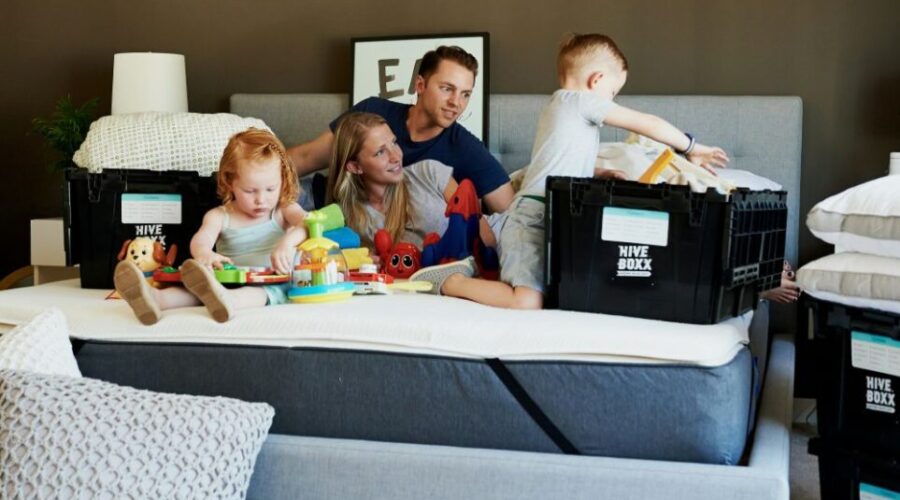 A couple and two young children sit on a bare king bed mattress with moving boxes positioned around them. A little girl plays with toys scattered around while the little boy pulls items out of one of the boxes. Parents lounge on bed between the children looking happily relaxed. This is how to make a house feel like a home: allow the time and space in the process to keep spirits high and relaxed