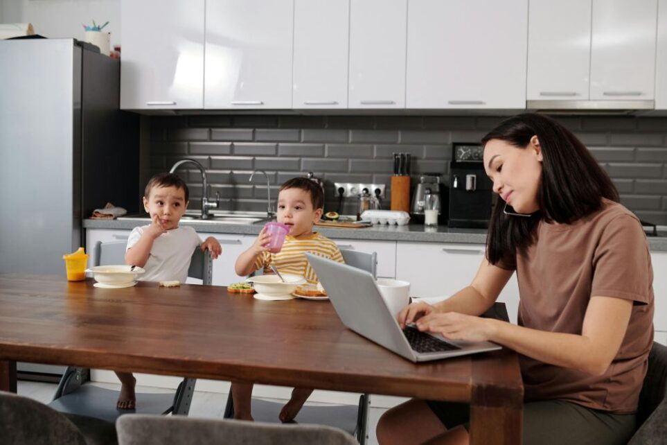 A busy mom sits at kitchen table while typing and on the phone and her two young boys sit at the table eating their lunch