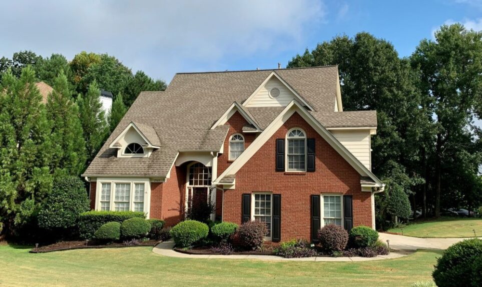 A basic large suburban brick house is shown on a mowed grass lot flanked by dense trees in the back and small bushes lining the front