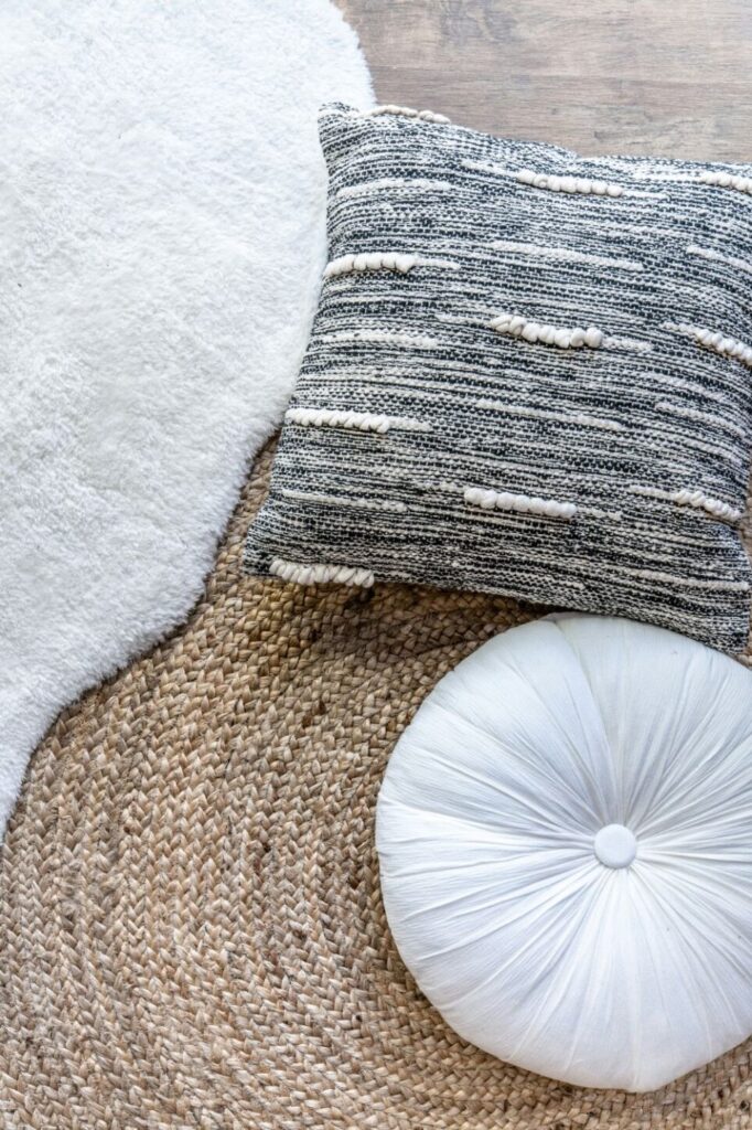 Top-down view of a textured gray and white square throw pillow, a tufted round white throw pillow, and a round-edged soft white rug over a round woven tan rug