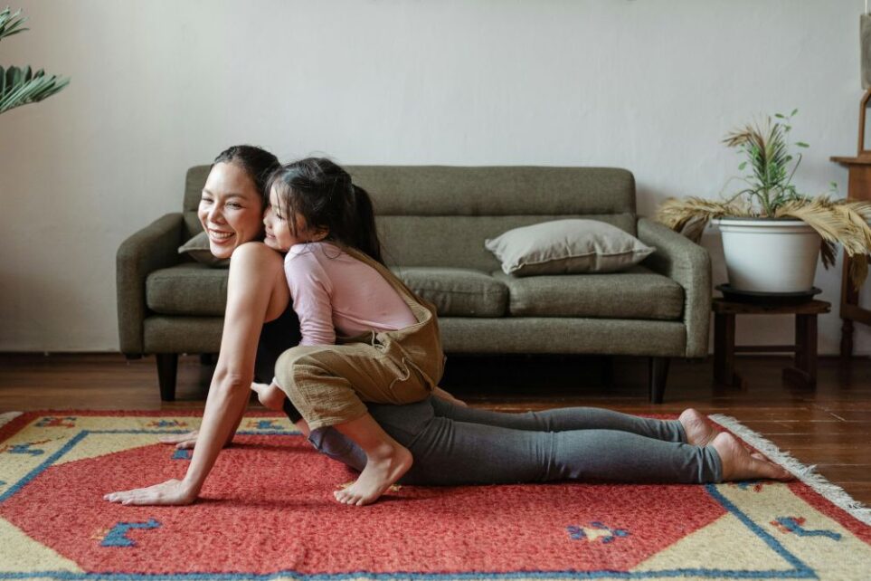 A smiling mom leans on hands with arms outstretched (upward-facing dog yoga pose) on living room rug with young daughter crouched on her back, happily playing together in living room