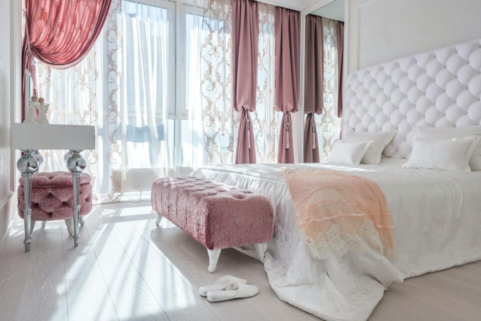 A beautiful and well-lit bedroom with a wall of windows and light, airy draperies furnished with a bed in white bedding and framed with a white plush upholstered headboard, a settee and matching stool in mauve velvet with matching mauve drapes, and an elegant vanity