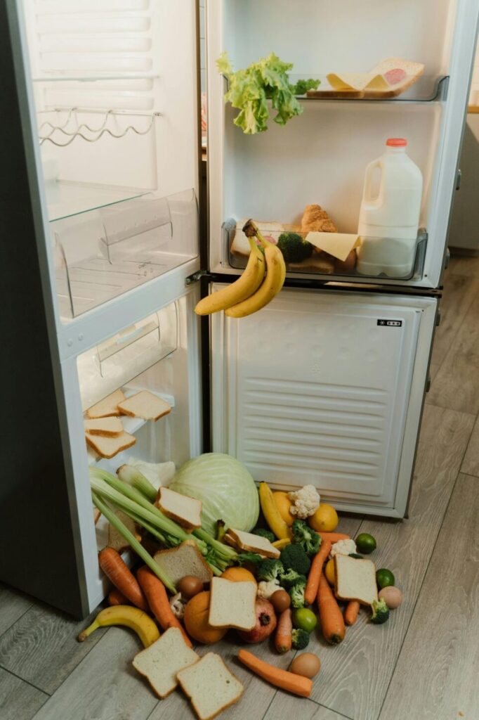 A refrigerator door is open to reveal an avalanche of produce and bread slices tumbling out of the bottom crisper drawer
