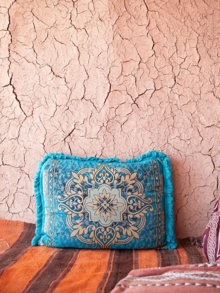 Rectangular turquoise throw pillow with gold detailing/design propped up over orange and brown striped rug against a cracked clay wall, a beautiful image of comfy and attractive floor seating
