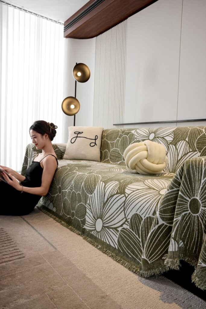 A couch is covered in a soft green and white throw blanket/covering, and a knotted cream coloured pillow along with a white square pillow rests on top. A woman sits contentedly on the floor leaning against the front of the couch and reading a book