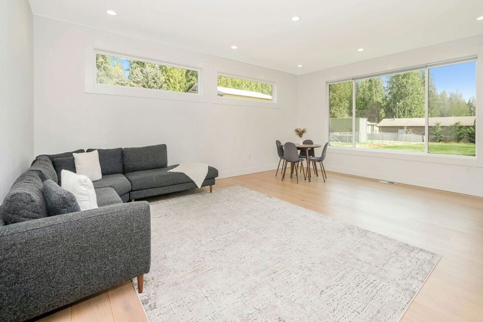 A neat but sparsely-furnished living room: a dark gray sectional couch and small round table with hard gray chairs are the only furnishings in an otherwise large and vacant space which, while well-lit and covered with a medium-sized gray area rug, is otherwise uninviting and uninspiring due to its unbalanced stuff/space ratio