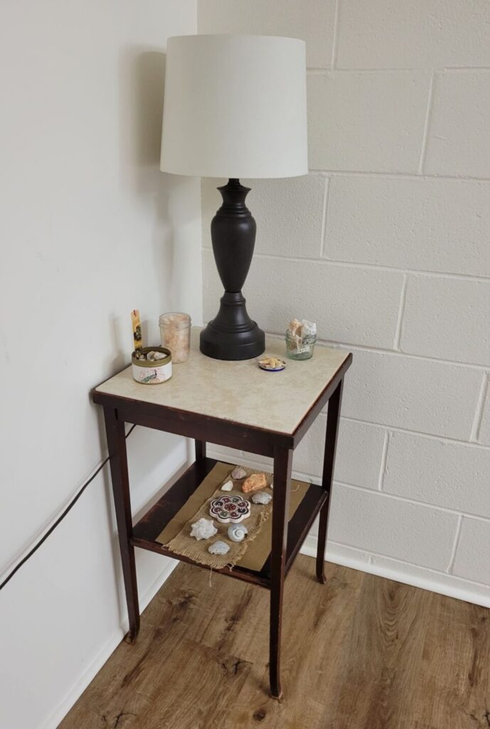 A small end table stands in a basement corner and holds a table lamp as well as other small decorations such as clear jars of seashells, salt crystals, and other natural trinkets