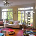 The sunroom: a white loveseat is positioned between two sides of floor-to-ceiling windows, with two end tables on either side and a tall open shelving unit to the right with more plants. Left of the left-side end-table (painted in multiple bright colours) and airplane plant on top is an heirloom rocking chair with a bright coral throw pillow. Two more bright throw pillows in other colours are on the loveseat. Along the right wall is a Pikler triangle and other toys, in the center of the room on the colourful area rug is a sensory spinning seat, and on the wall opposite the windows is a child's blue desk and chair for schoolwork. Partially in the picture to the left is a small trampoline for sensory breaks, and along the left wall are more windows (and more plants out of sight). Plenty of sunlight, colours, plants, and sensory toys/furniture, as here in the sunroom, are all optimal features for creative spaces