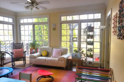 The sunroom: a white loveseat is positioned between two sides of floor-to-ceiling windows, with two end tables on either side and a tall open shelving unit to the right with more plants. Left of the left-side end-table (painted in multiple bright colours) and airplane plant on top is an heirloom rocking chair with a bright coral throw pillow. Two more bright throw pillows in other colours are on the loveseat. Along the right wall is a Pikler triangle and other toys, in the center of the room on the colourful area rug is a sensory spinning seat, and on the wall opposite the windows is a child's blue desk and chair for schoolwork. Partially in the picture to the left is a small trampoline for sensory breaks, and along the left wall are more windows (and more plants out of sight). Plenty of sunlight, colours, plants, and sensory toys/furniture, as here in the sunroom, are all optimal features for creative spaces