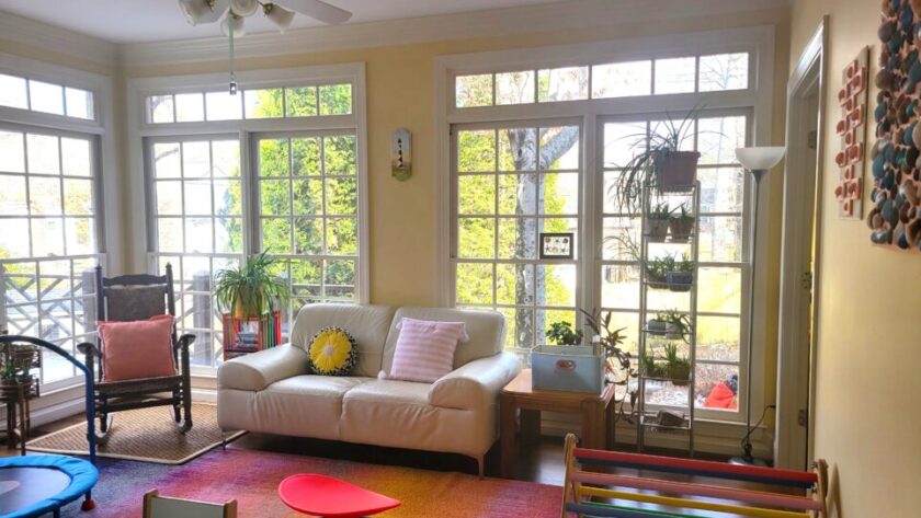 The sunroom: a white loveseat is positioned between two sides of floor-to-ceiling windows, with two end tables on either side and a tall open shelving unit to the right with more plants. Left of the left-side end-table (painted in multiple bright colours) and airplane plant on top is an heirloom rocking chair with a bright coral throw pillow. Two more bright throw pillows in other colours are on the loveseat. Along the right wall is a Pikler triangle and other toys, in the center of the room on the colourful area rug is a sensory spinning seat, and on the wall opposite the windows is a child's blue desk and chair for schoolwork. Partially in the picture to the left is a small trampoline for sensory breaks, and along the left wall are more windows (and more plants out of sight). Plenty of sunlight, colours, plants, and sensory toys/furniture, as here in the sunroom, are all optimal features for creative spaces
