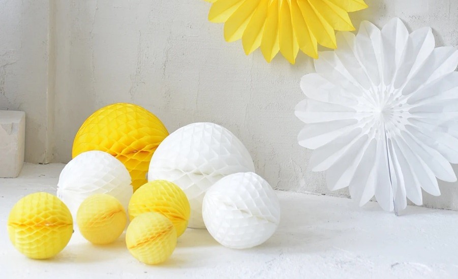 White and yellow paper honeycomb pompoms of various sizes rest on the floor in front of a white stucco wall where a large white paper fan stands and part of a yellow paper fan is seen hung above it