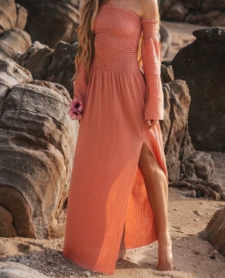 Close-up photo of salmon pink off-shoulder linen maxi dress as seen on model. Loose and flowy, a slit goes just above knee for a breezy style. Long sleeves hang down arms from bare shoulders.